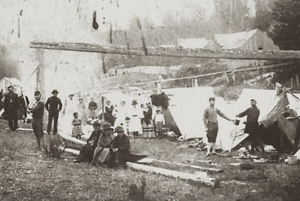 A vintage photograph of a group of people, including Indigenous individuals, gathered at the Old Man House in Suquamish, WA, with tents and early structures in the background.