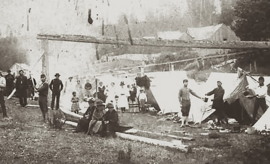 A vintage photograph of a group of people, including Indigenous individuals, gathered at the Old Man House in Suquamish, WA, with tents and early structures in the background.