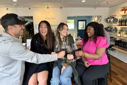 Group of friends enjoying wine at Eleven Winery, smiling and toasting with glasses of red wine. The relaxed and cheerful atmosphere inside the tasting room includes shelves with wine bottles in the background, highlighting a welcoming and social experience.