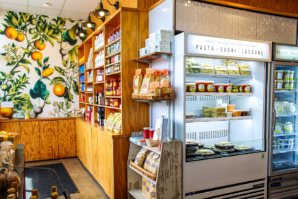 Interior of Via Rosa 11 on Bainbridge Island, featuring a cozy display of Italian goods including shelves stocked with pasta, sauces, and other specialty products. A brightly decorated wall with citrus artwork and refrigerated sections displaying fresh pasta and lasagna add to the authentic Italian deli atmosphere.