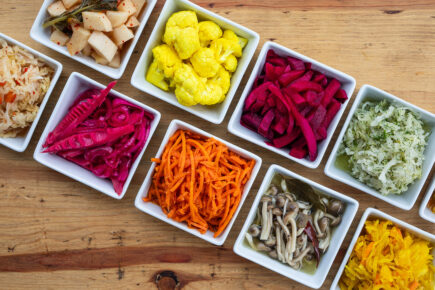 A colorful assortment of fermented vegetables from Iggy's Alive & Cultured, displayed in small square dishes on a wooden table. The vibrant selection includes pickled cauliflower, shredded carrots, various types of kimchi, beets, mushrooms, and other fermented produce, showcasing a variety of textures and flavors.