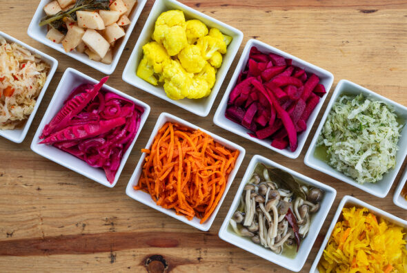 A colorful assortment of fermented vegetables from Iggy's Alive & Cultured, displayed in small square dishes on a wooden table. The vibrant selection includes pickled cauliflower, shredded carrots, various types of kimchi, beets, mushrooms, and other fermented produce, showcasing a variety of textures and flavors.