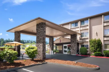 Exterior view of Quality Inn & Suites Silverdale with covered entrance, stone columns, landscaped patio seating, and clear blue sky.