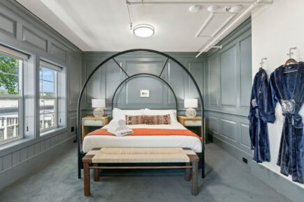 Elegant hotel room at Hotel Scandi featuring a large bed with an arch canopy, vibrant orange blanket, and decorative pillows, flanked by stylish nightstands with lamps. The room has paneled gray walls, large windows with natural light, a bench at the foot of the bed, and plush robes hanging on the wall.