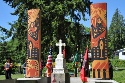 Chief Seattle's gravesite with two vibrant totem poles, honoring the Suquamish leader. The American flag and other ceremonial flags are placed around the site, surrounded by lush greenery.