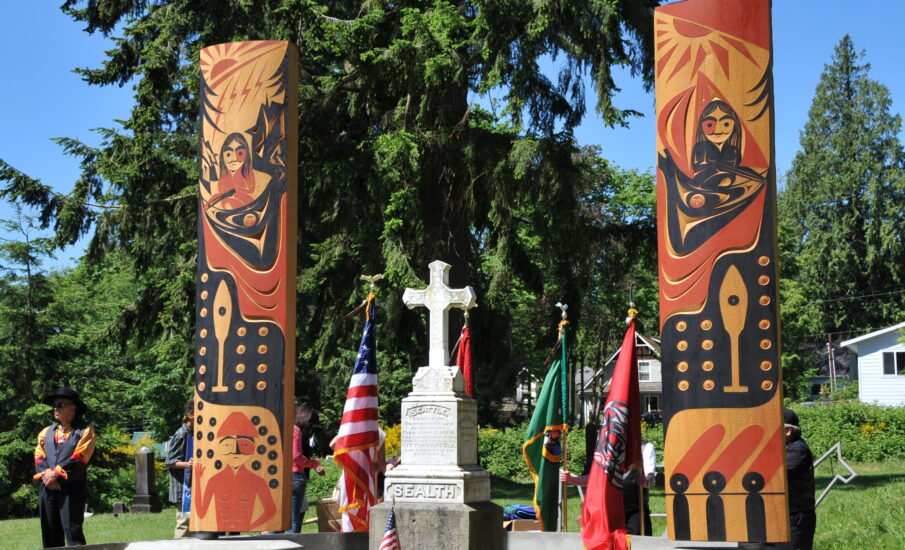 Chief Seattle's gravesite with two vibrant totem poles, honoring the Suquamish leader. The American flag and other ceremonial flags are placed around the site, surrounded by lush greenery.