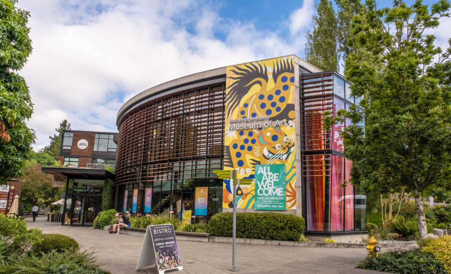 A modern, circular building with a wooden slatted facade and large glass windows houses the Bainbridge Island Museum of Art. A colorful banner featuring a stylized mythical creature and the words "ALL ARE WELCOME" hangs prominently on the exterior. The entrance is flanked by greenery, with a sidewalk leading to a bistro sign in the foreground. The museum is located in a lively area with visitors sitting outside and walking nearby, and the sky is partly cloudy.