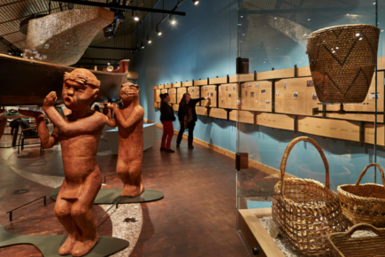 An interior view of the Suquamish Museum, showcasing intricately carved wooden figures holding a canoe, woven baskets, and historical information panels lining the wall.