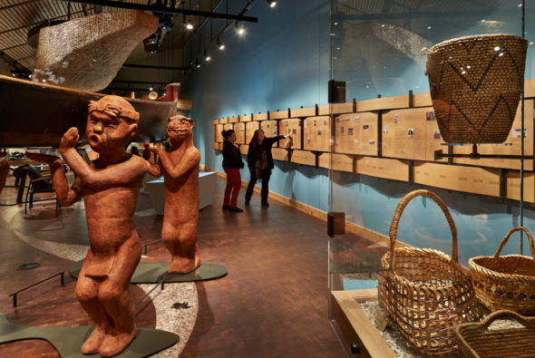 An interior view of the Suquamish Museum, showcasing intricately carved wooden figures holding a canoe, woven baskets, and historical information panels lining the wall.