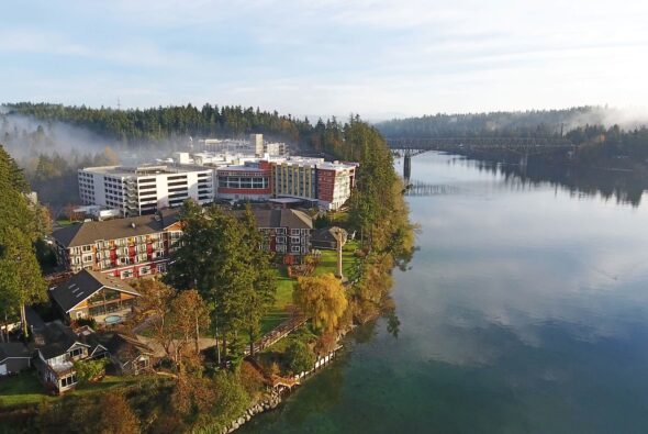 An aerial shot of the Clearwater Casino & Resort property shows a beautiful waterfront landscape. The main resort building stands prominently with colorful balconies, surrounded by lush greenery and smaller structures. The calm water of the inlet reflects the serene surroundings, and a bridge is visible in the distance, spanning the water. Light mist gently floats above the evergreen forest, enhancing the peaceful and picturesque atmosphere of this Pacific Northwest destination.