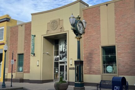 The photo shows the front entrance of the Kitsap History Museum in Bremerton, WA. The building has a classic, brick-and-tan facade with a sign reading "Museum" next to the entrance. Above the door is an emblem representing Kitsap's heritage, while large windows feature historical images. In front of the museum is a decorative clock post with ornate black and gold detailing, and a blue USPS mailbox sits nearby. The scene is calm and inviting, with clear skies above, creating a welcoming atmosphere for visitors.