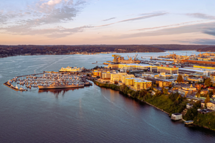 Aerial view of Bremerton's waterfront, featuring the marina with several docked boats, naval ships, and surrounding residential and commercial buildings. The landscape is bordered by a calm, reflective bay, with the distant horizon revealing lush, forested areas and a sunset-lit sky. Naval shipyards and industrial facilities are visible in the background, adding contrast to the natural scenery and harbor area. The image captures Bremerton’s unique mix of maritime activity, urban development, and natural beauty, all set against the Kitsap Peninsula's coastal landscape.