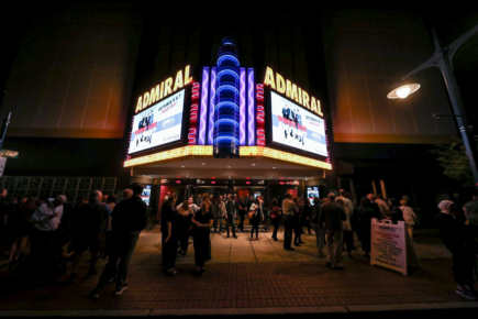 Nighttime exterior of the Admiral Theatre in Bremerton, Washington, illuminated with vibrant marquee lights displaying upcoming shows. A lively crowd gathers on the sidewalk, enjoying the bustling atmosphere of the historic venue.