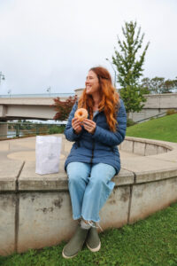 Emma sitting on a concrete bench, smiling while holding a donut from Larry & Kristi’s Scratch Bakery, with a paper bag next to her, at a park in Bremerton, WA.