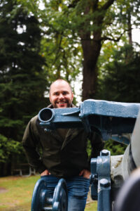 Emma’s husband smiling as he stands behind a cannon at Illahee State Park, surrounded by tall green trees.