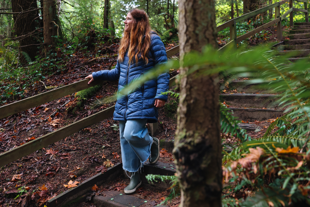 A woman wearing a blue puffer jacket and rain boots walks down a forest trail surrounded by tall trees and lush greenery, smiling as she enjoys the peaceful atmosphere of the woods.