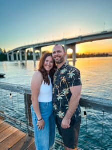 Emma and her husband smiling while standing on the deck at The Boat Shed in Manette, WA, with a stunning sunset and the Manette Bridge in the background, overlooking the water.