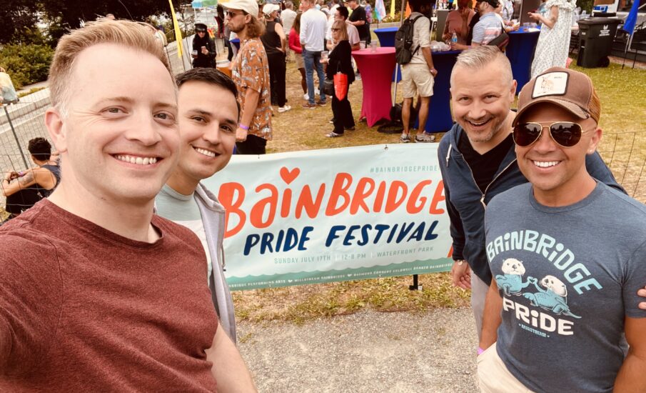 A group of four friends smiling at the camera during the Bainbridge Pride Festival. On the left are Kenneth Minogue and Erik Axthelm, and on the right are Christopher Lindstrom and Joey Chapman. Behind them is a colorful festival scene with people enjoying the event, vibrant umbrellas, and a banner that reads 'Bainbridge Pride Festival, Sunday, July 17th, 12-8 PM, Waterfront Park.