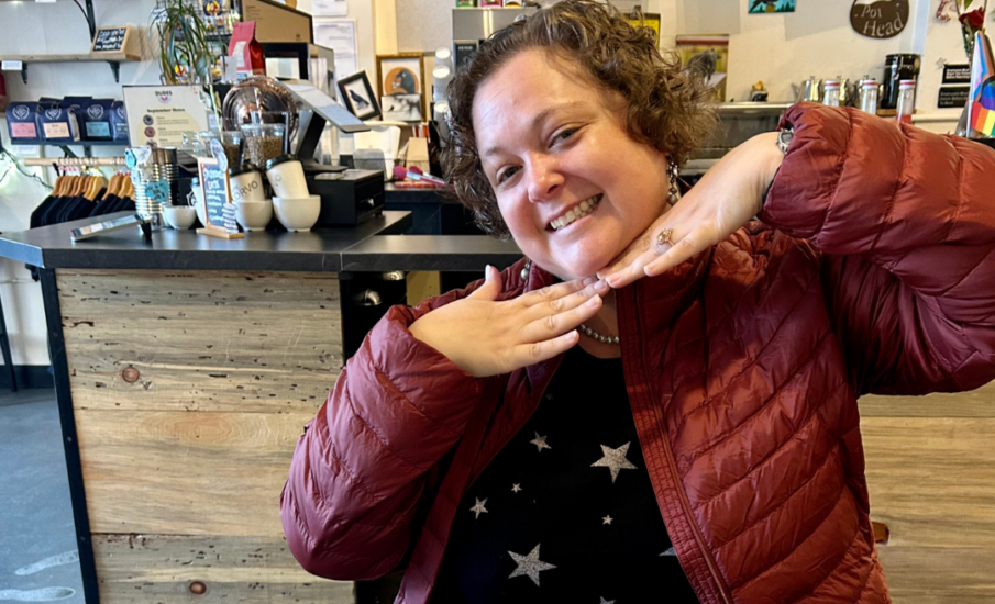 Jess Senires, smiling brightly in a cozy coffee shop, wearing a red puffy jacket and a star-patterned shirt. She’s seated at a table with a cappuccino and a sprinkled donut, hands posed playfully under her chin.