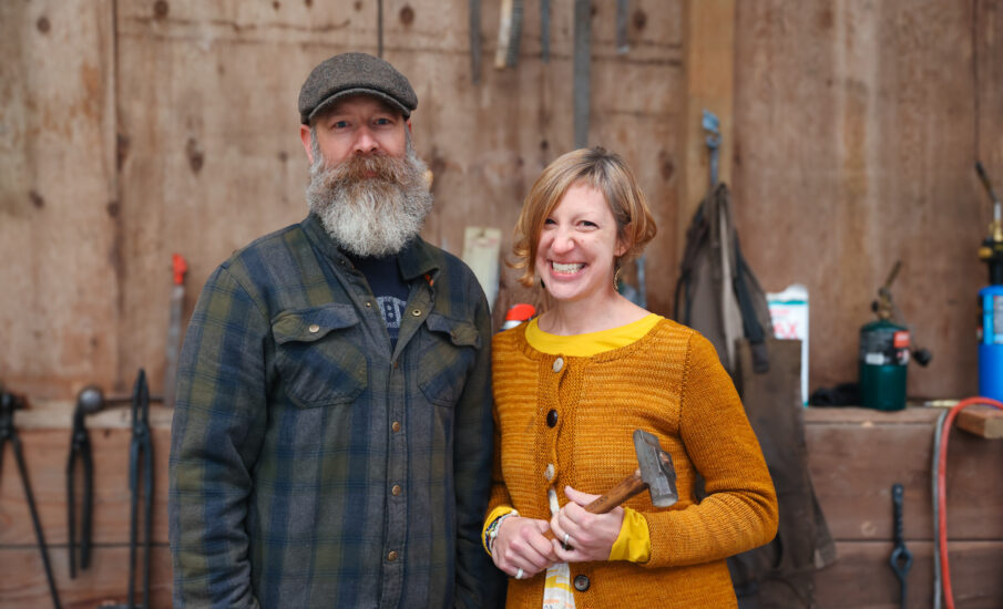 A couple standing in a workshop setting. The man has a bushy beard and is wearing a plaid shirt, while the woman is dressed in a mustard-colored sweater and holding a small hammer. The background features tools hanging on a wooden wall.