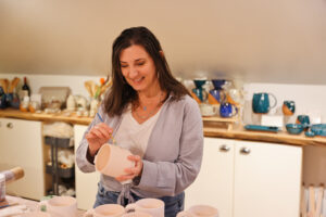 Meredith Chernick, the artist behind Fern Street Pottery, carefully paints a ceramic cup while standing in her cozy, well-lit studio filled with pottery.