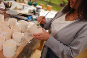 A close-up of Meredith painting a green design onto a pottery cup, with a table full of ceramic pieces and tools visible in the background.