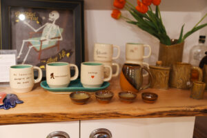 A collection of handmade mugs, bowls, and other ceramic items displayed on a wooden shelf, with an art print in the background and a vase of tulips for decoration.