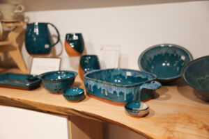 An assortment of blue-glazed pottery pieces, including bowls, a pitcher, and serving dishes, arranged on a wooden shelf, showcasing Meredith's distinctive style.