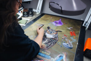 A female artist with glasses works intently at her desk, painting an elaborate scene that includes an owl and various nature-themed elements. The studio space is filled with art supplies, and a window provides a view of greenery outside.