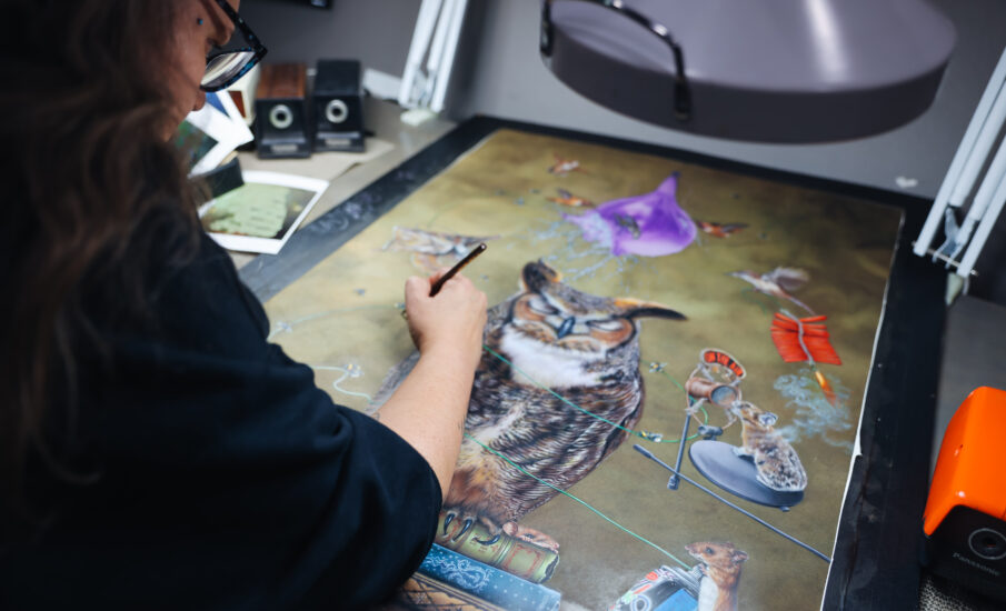 A female artist with glasses works intently at her desk, painting an elaborate scene that includes an owl and various nature-themed elements. The studio space is filled with art supplies, and a window provides a view of greenery outside.