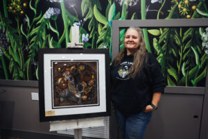 An artist standing beside a framed artwork depicting a nature-inspired still life. The background is decorated with a mural of green leaves and white flowers, adding a vibrant touch to the studio environment.