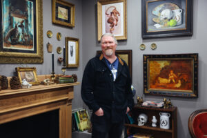 A male artist stands in his studio, surrounded by framed artworks, antiques, and natural objects like pinecones and animal skulls. The eclectic décor gives the space a vintage and whimsical atmosphere.