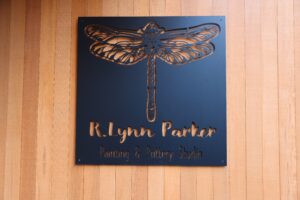 A black metal sign for R. Lynn Parker Painting & Pottery Studio, featuring an intricate cutout of a dragonfly. The sign is mounted on a wooden wall, creating a natural and rustic look.