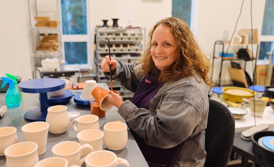 Rebecca Parker, the artist, smiling while painting a ceramic mug at her worktable, is surrounded by pottery pieces and supplies. The studio is brightly lit, with shelves and pottery tools in the background.