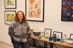 Rebecca Parker, the artist standing beside a display of pottery, with framed abstract artwork hanging on the wall behind her. She is smiling and appears to be in a gallery or studio setting.