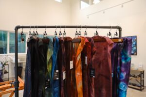 A rack displaying hand-dyed silk scarves in a range of rich colors, from deep greens to vibrant oranges and purples. The scarves hang neatly in a well-lit studio space.