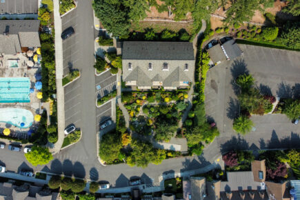 Aerial view of The Inn at Pleasant Beach, showcasing the layout of the property with landscaped gardens, a swimming pool area, parking spaces, and surrounding buildings. The scene captures the inviting outdoor pool, seating areas with umbrellas, and the lush greenery that surrounds the inn, creating a welcoming and serene atmosphere.