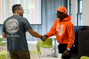 Two men are shaking hands at Creative Mornings Poulsbo. One is wearing an orange hoodie with a message that reads, "Me vs Me, I am my competition," while the other wears a gray t-shirt featuring "The Ship Inn" graphic. They are standing in a bright room with green chairs, with a colorful screen displaying the Creative Mornings logo in the background, conveying a sense of community and collaboration.