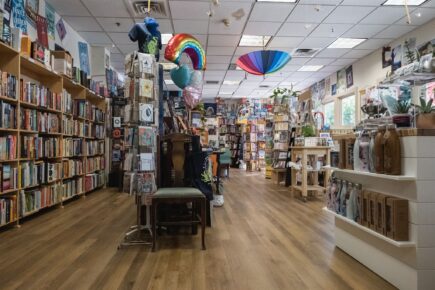 Interior of Ballast Book Company, showcasing rows of bookshelves filled with a variety of books, alongside displays of greeting cards, gifts, and unique items. The shop has a welcoming atmosphere with colorful decorations like hanging umbrellas and balloons, creating a cozy and whimsical browsing experience.