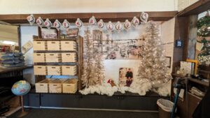 Holiday-themed display at a store featuring a banner that says 'Happy Holidays,' decorated silver Christmas trees, and historical photos of the town, with crates stacked on shelves and a vintage globe on the left.