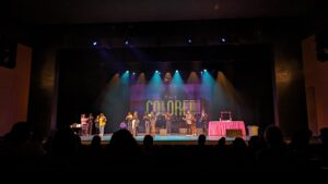 A group of performers takes a bow on stage at the end of a production titled "The Colored Museum." The audience is silhouetted in the foreground, watching the cast under bright stage lights.