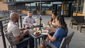 Four friends enjoying a meal together on an outdoor deck at a restaurant, with a beautiful view of the water and mountains in the background. The group is seated at a table, sharing food and drinks in a relaxed, social atmosphere.