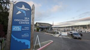 A signboard at the Sea Discovery Center in Poulsbo, WA, with a logo of an orca. The sign shows the public aquarium's opening hours, located near a marina, with the building in the background under a clear sky.