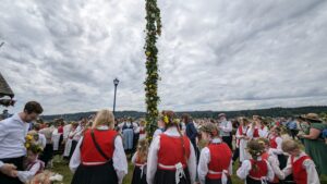 A midsummer celebration in Poulsbo, WA, with a large maypole adorned with flowers and participants dressed in traditional Scandinavian attire. The gathering includes adults and children with floral headpieces, enjoying the festive atmosphere under a cloudy sky.