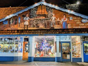 The exterior of Sluys Bakery and Indigo Plum store in Poulsbo, WA, illuminated at night. The bakery's facade features a mural depicting bakers working with a large stone oven, with one person dressed in traditional Norwegian attire. Above the mural are the words "Give us this day our daily bread" alongside the bakery's logo. Below the mural, the storefront displays warm lighting and a festive window display featuring various items, while the Indigo Plum store next door shows a brightly lit interior filled with shoes and clothing.