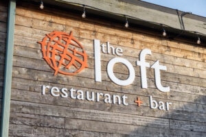 The exterior sign for "the loft restaurant + bar" mounted on a rustic wooden wall, featuring an orange circular logo with abstract lines next to the restaurant's name. The sign is illuminated by small string lights above it.