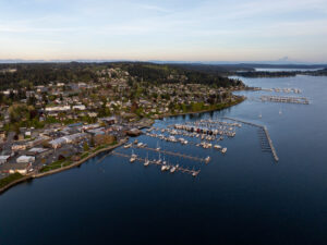 Aerial view of Poulsbo, WA, at sunset with the marina in the foreground, filled with boats and yachts. The serene waters of Liberty Bay reflect the golden hues of the setting sun, while the lush, forested landscape stretches across the background. Snow-capped peaks of the Olympic Mountains can be seen under a partially cloudy sky, creating a picturesque Pacific Northwest scene.