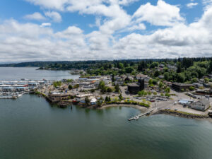 An aerial view of Poulsbo, WA, showcasing the waterfront with boats docked at the marina, surrounded by lush greenery under a partly cloudy sky.