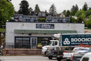 The front view of Peninsula Beverage Co. in Poulsbo, with the building's sign visible and a delivery truck parked in front, featuring a green, forested background.