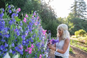 A woman with light blond hair enjoys picking vibrant sweet pea flowers in a lush garden at Farm Kitchen. She is holding a small bouquet and appears to be admiring the flowers' fragrance. The scene is surrounded by colorful blossoms in shades of pink, purple, and white, set against a backdrop of greenery and soft sunlight filtering through the trees.